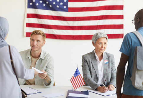 Election workers assist voters at a registration table with a U.S. flag behind them